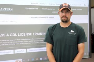 A man stands with his back against a digital presentation board showing a CDL training program website.