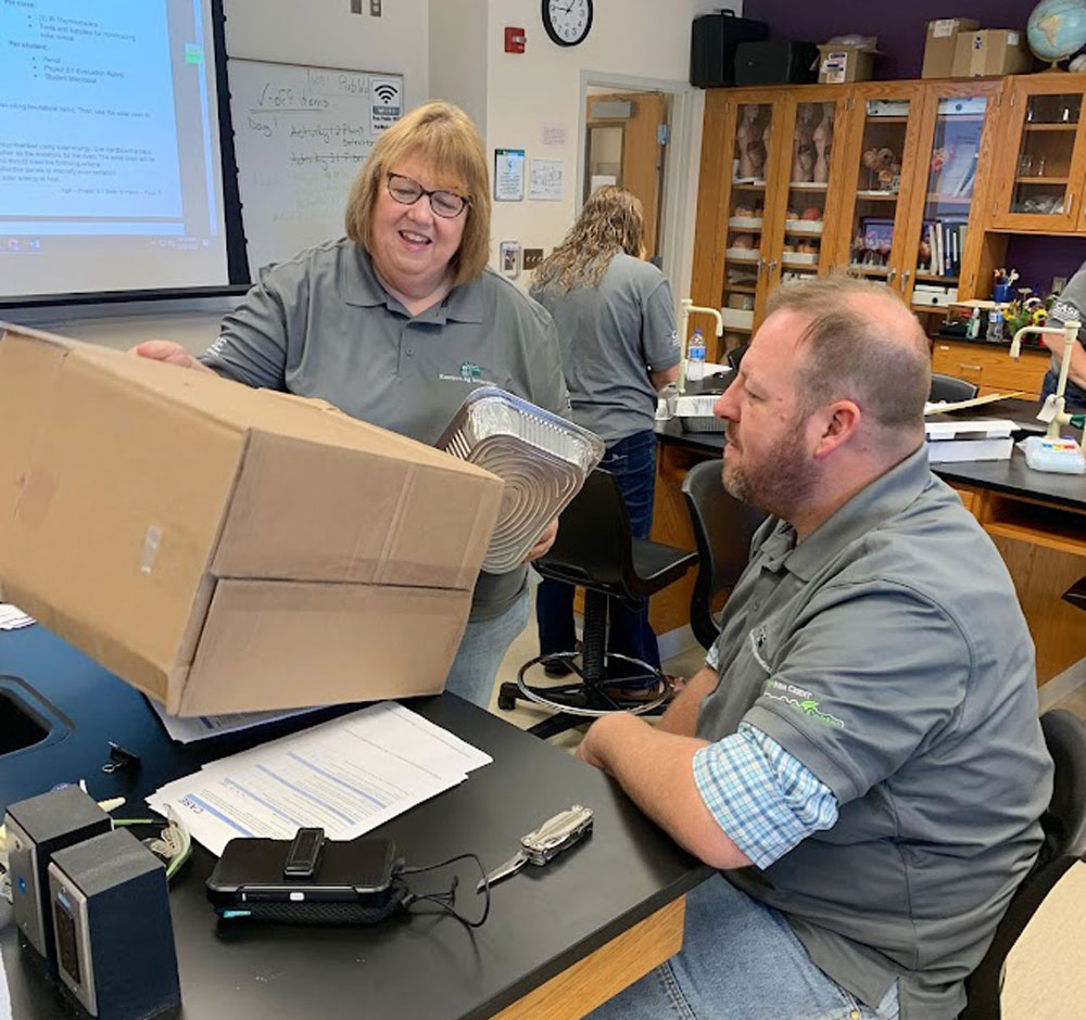 Two teachers look at a cardboard box, part of a lab experiment to cook using the heat of the sun.