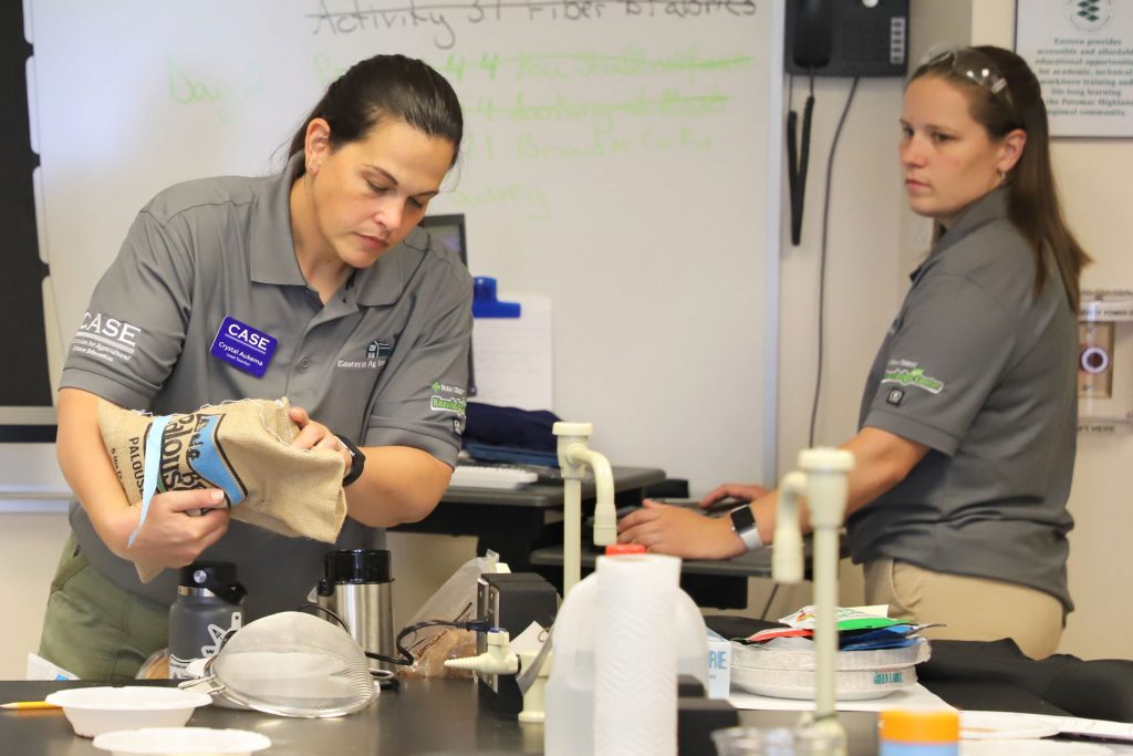 Two teachers in front of laboratory classroom, with one pouring wheat into a coffee grinder for an experiment.