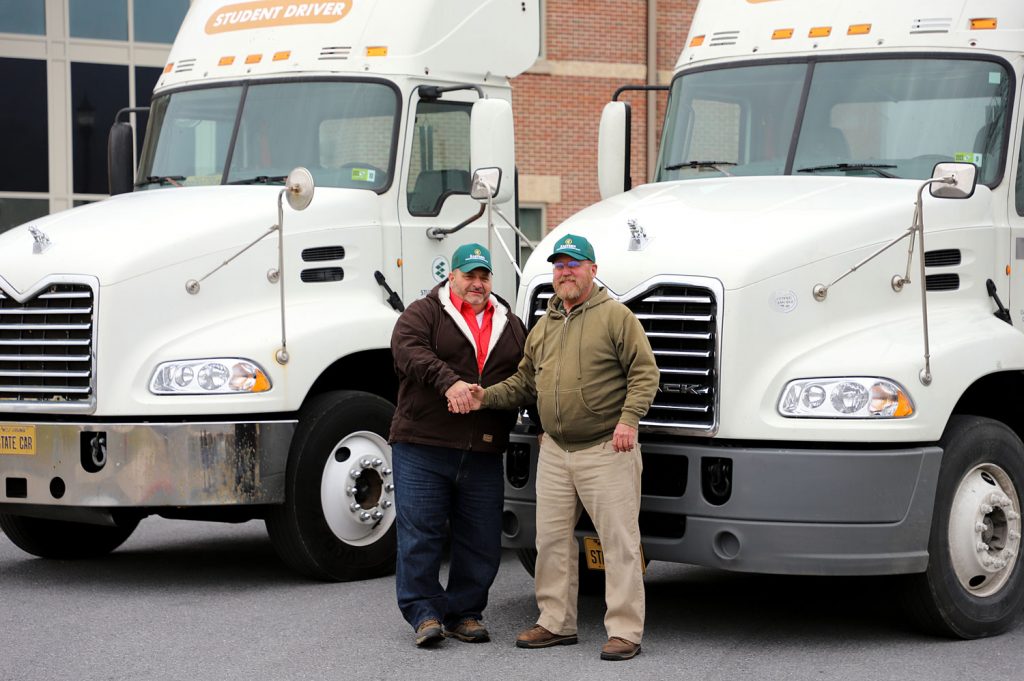Two men stand in front of two tractor trailers and shake hands.