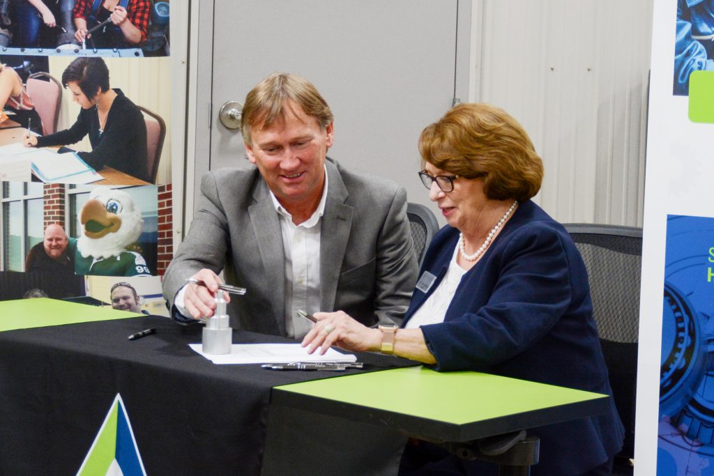 Two people sit next to one another a a desk signing a piece of paper. The two are the presidents of two community colleges, and they are agreeing to a partnership so students of one school can take classes at the other school's locations.