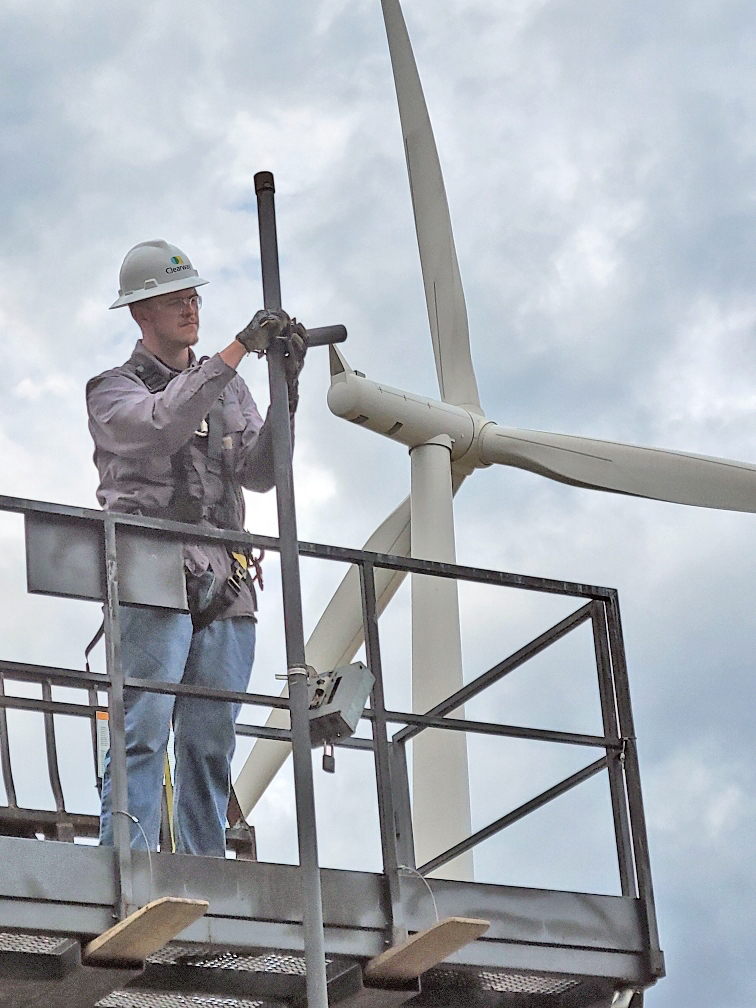 A man wearing a hard hat works on a platform. A wind turbine is behind him.