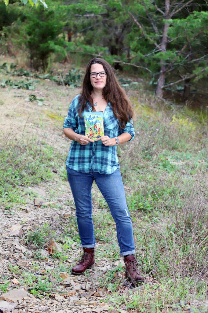 A woman stands on a rocky path. She is holding a book. The cover of the book says, "The Hike to Home."