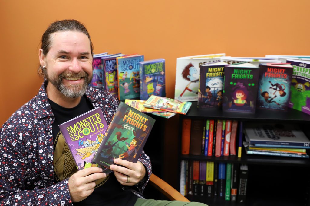 A man displays two books in his hands. A bookshelf holding many other books is behind him.