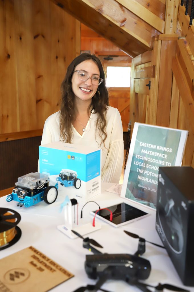 A woman wearing glasses stands behind a table with several technology-related items on display.