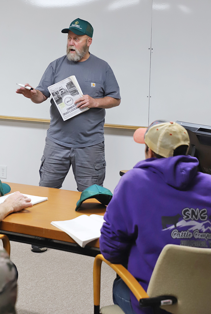 A man stands in front of a classroom, holding a manual in one hand, and gestures toward the students while holding a whiteboard marker with the other. 