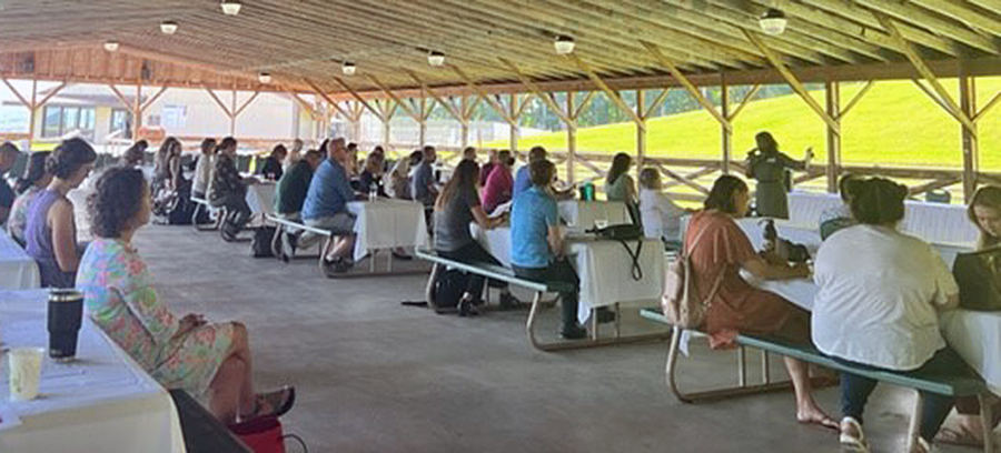 A group of people sitting on picnic tables in an open-air shelter listen to a speaker in the front of the group.