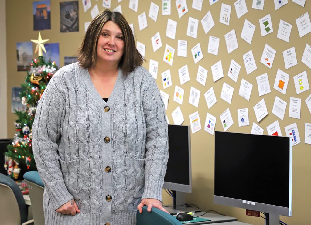 A woman stands next to a couple of computers in a classroom.