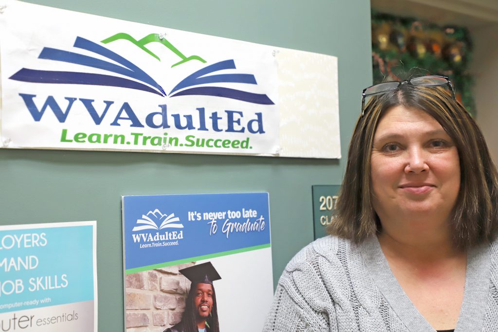 A woman stands next to the door of a classroom with a sign "WVAdultEd" displayed on the wall next to her.
