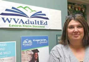 A woman stands next to the door of a classroom with a sign "WVAdultEd" displayed on the wall next to her.